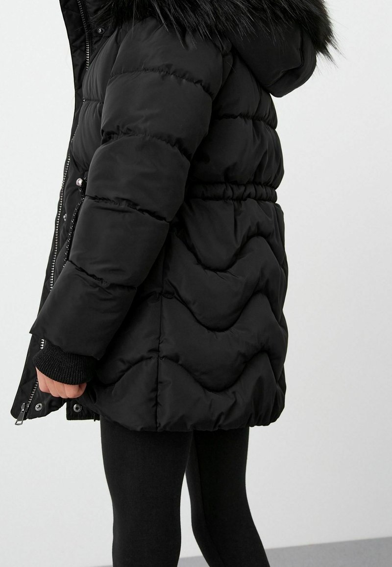 Personne portant un manteau d'hiver matelassé noir avec une capuche bordée de fourrure, une fermeture éclair et des boutons-pression, associée à des leggings noirs sur un fond blanc.