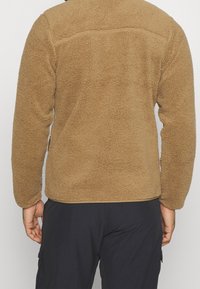 Brun fleecejacka med texturerad yta, långärmad och med rak nederkant, bärs över svarta byxor. Bakifrån syns en sömlös design.