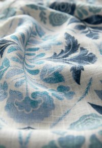 Tissu blanc avec motifs floraux bleus et turquoise, montrant un tissage texturé et des plis doux.