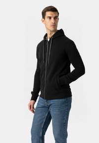 Črna jopa z zadrgo in kapuco z vrvico, rebrastimi manšetami in stranskimi žepi, združena z modrimi jeans hlačami iz denima. Tkanina izgleda gladka.