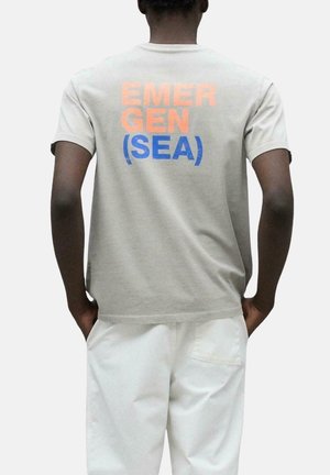 Camiseta de algodón gris con mangas cortas, con el texto naranja y azul "EMERGEN (SEA)" impreso en la parte posterior. Diseño sencillo y de corte relajado.