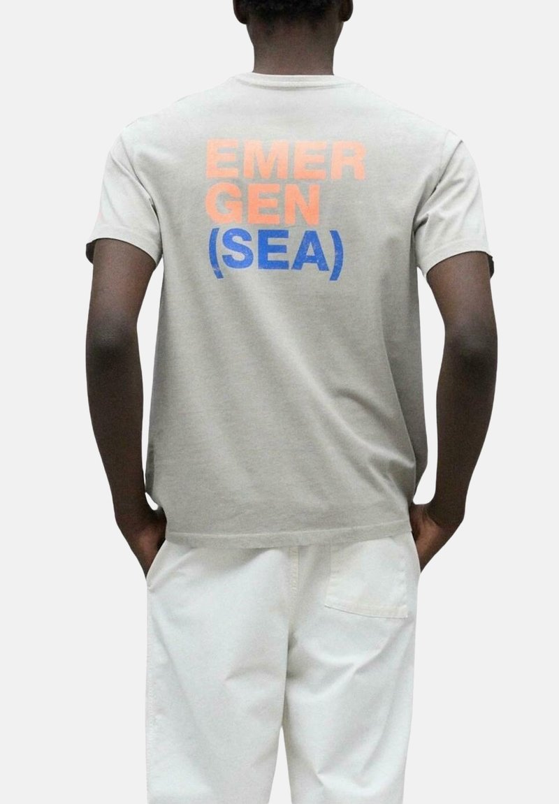 Camiseta de algodón gris con mangas cortas, con el texto naranja y azul "EMERGEN (SEA)" impreso en la parte posterior. Diseño sencillo y de corte relajado.