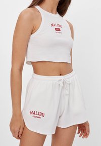 Bílý žebrovaný crop top a odpovídající šortky, na kterých je červený text "MALIBU". Top má vysoký výstřih a šortky mají elastický pas.