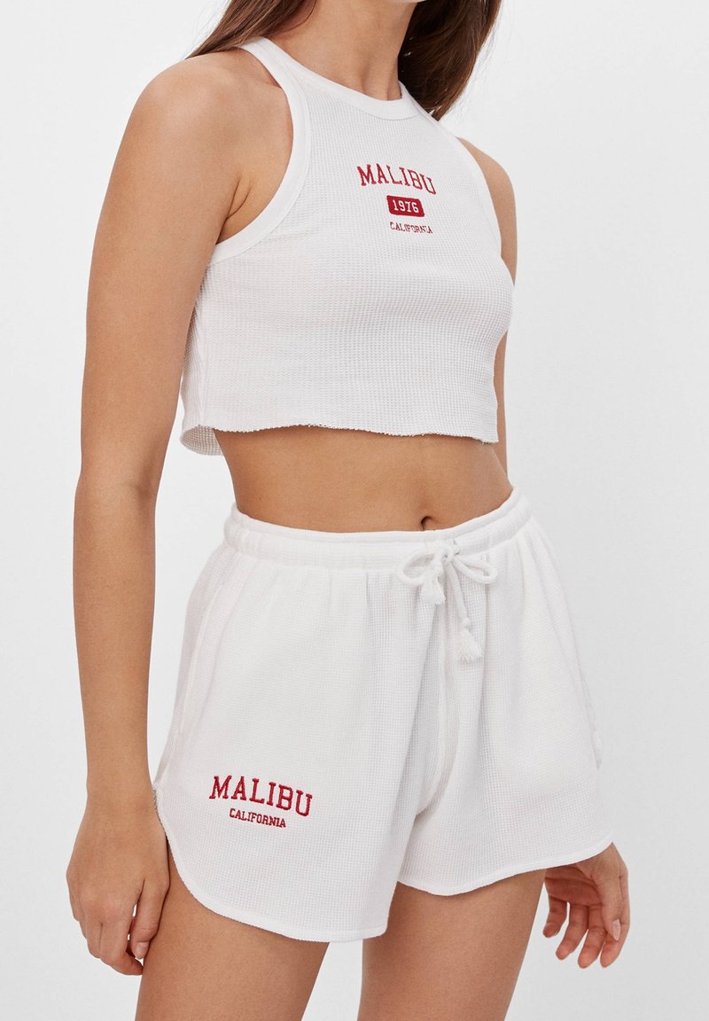 Bílý žebrovaný crop top a odpovídající šortky, na kterých je červený text "MALIBU". Top má vysoký výstřih a šortky mají elastický pas.