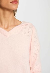 Maglione rosa lavorato a maglia con scollo a V e cappuccio. Presenta motivi geometrici traforati sulle maniche, con una texture morbida e pelosa.
