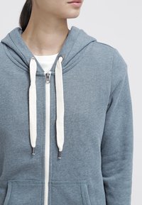 Felpa con cappuccio e zip di colore blu chiaro, realizzata in un tessuto morbido. Presentatringenti beige, tasche frontali e un taglio casual.