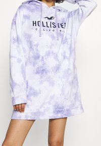 Fialová tie-dye mikinová šaty s přední kapsou, kapucí se šňůrkami a černým logem "Hollister California" na hrudi. Měkký a pohodlný materiál.