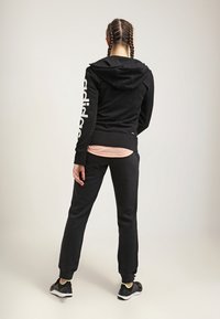 Ensemble de sport noir avec capuche, arborant le logo blanc "adidas" sur la manche. Pantalons ajustés et haut près du corps, fabriqués dans un matériau doux.