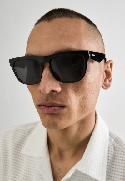 Gafas de sol rectangulares negras con lentes oscuros, que presentan un marco grueso, un diseño geométrico y sutiles acentos metálicos en las patillas.
