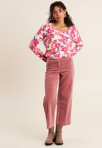 Blumenmuster-Pullover in pinken, roten und lila Farbtönen. Kombiniert mit weiten, rosafarbenen Cordhosen mit seitlichen Knöpfen und glatter Textur.