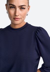 Temno moder top s kratkimi napihnjenimi rokavi, okrolim izrezom in gladko teksturo tkanine. Ima minimalno šivanje in čist dizajn.