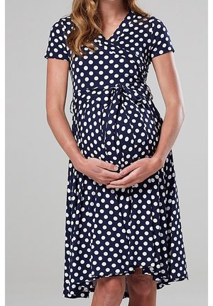 Raskaana oleva nainen pukeutuneena laivastonsiniseen pilkulliseen mekkoon, kädet hellästi tukemassa kasvavaa vauvavatsaa neutraalia harmaata taustaa vasten.