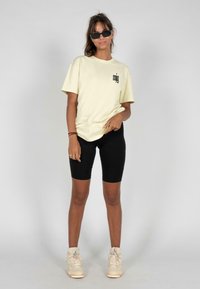 Lys gul bomulds t-shirt med sort grafik, parret med sorte shorts. Båret med beige sneakers og sorte rektangulære solbriller.