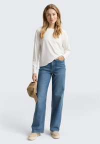Vrouw in een witte lange mouwen top, blauwe wijde jeans, beige laarzen, die een bruine pet vasthoudt, staand tegen een effen lichte achtergrond.