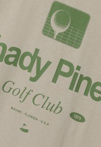 Crèmekleurige stof met groene tekst: "Shady Pines Golf Club," een logo van een golfbal, en details zoals "1979" en "Miami, Florida, VS."