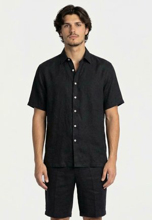 Uomo che indossa una camicia nera a maniche corte con bottoni e pantaloncini neri abbinati, in piedi davanti a uno sfondo bianco uniforme.