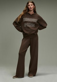 Brązowa bluza z kapturem z napisem "BISOUS PARIS", w zestawie z szerokimi spodniami. Miękka tkanina, luźny krój i minimalistyczny design.