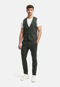 Dunkelgrüne Weste über einem weißen T-Shirt, kombiniert mit passenden dunklen Hosen und weißen Sneakers. Glatter Stoff, moderne Passform, minimalistische Details.