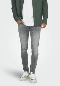 Jeans in denim grigio con vestibilità slim, design sbiadito e dettagli di sfumatura leggeri. Abbinati a una t-shirt bianca e una camicia verde a bottoni.