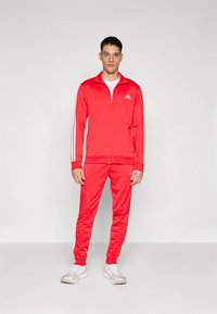 Fato de treino atlético vermelho com detalhes brancos, composto por um casaco com fecho éclair e calças a condizer, feito de tecido liso e elástico.