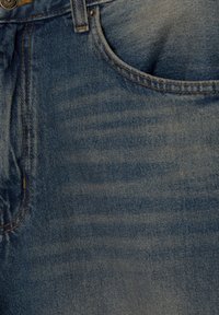 Gros plan sur la poche d'un jean en denim bleu avec des coutures visibles, une passants de ceinture et un détail de bouton en métal.