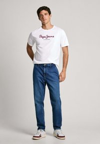 Bijela majica s logom "Pepe Jeans London" u burgundijskoj boji, uparena s plavim trapericama. Sportske tenisice upotpunjuju odjeću.