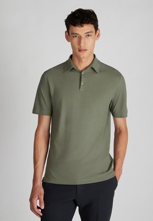 Jonge man in een olijfgroene polo met korte mouwen en twee knopen, staande tegen een effen lichtgrijze achtergrond.