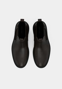 Pár tmavě hnědých kožených slip-on bot s černými podrážkami, kulatými špičkami a poutky na bílém pozadí.