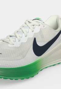 Witte Nike hardloopschoen met zwart swoosh-logo en groene zoolverloop, met witte veters en een gestructureerd bovenmateriaal.