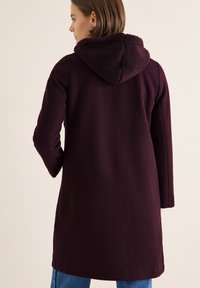 Lange bordeauxrode jas met een gestructureerde, pluche capuchon, voorzien van lange mouwen en een rechte snit, gecombineerd met blauwe jeans.
