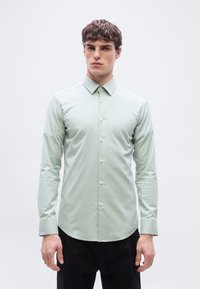 Νέος άντρας που φορά ένα ανοιχτό πράσινο πουκάμισο με κουμπιά και γιακά και μαύρο παντελόνι, στέκεται μπροστά σε απλό λευκό φόντο.