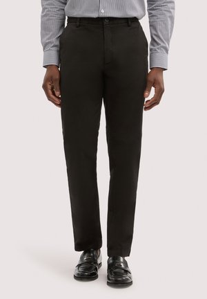 Mann trägt schwarze Stoffhose, schwarze Ledermokassins und ein gestreiftes Langarmshirt, steht frontal zur Kamera.