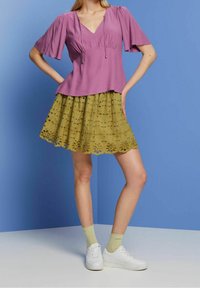 Top lilla a scollo a V con maniche corte a volants, abbinato a una gonna a strati verde oliva con ritagli floreali e sneakers bianche.