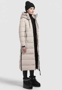 Long beige puffer coat with a hood, featuring quilted sections and black zippers. Worn over a black top and paired with black boots.