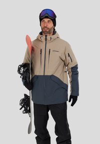 Sjezdová bunda v béžové a námořnické barvě, s zipy, kapucí, několika kapsami a texturovanou tkaninou, v kombinaci s černými lyžařskými kalhotami a výbavou.