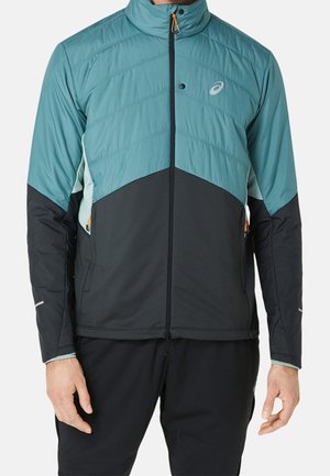Homme portant une veste zippée bleu sarcelle et noire avec un col montant et des manches longues, debout les bras détendus le long du corps.