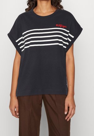 Kvinde iført sort kortærmet skjorte med hvide vandrette striber og rød "Hilfiger" tekst, kombineret med brune bukser.