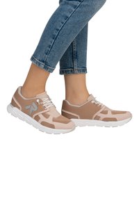 Baskets de sport beige et marron clair avec une tige en maille et une semelle en caoutchouc blanc, présentant un détail de logo et des accents contrastants.