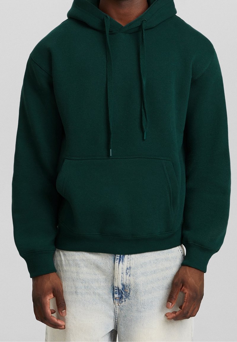 Grön huvtröja i tjockt material, med en framficka, snoddar och ribbade ärmslut och nederkant. Bärs med ljusblå jeans.