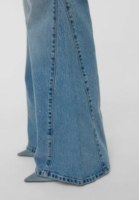 Benen in wijde blauwe denim jeans die gedeeltelijk de puntige grijze hoge hakken bedekken, tegen een effen achtergrond.