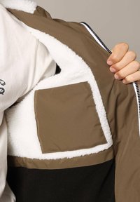 Bruine en zwarte ritssluiting jas met een witte fleece voering; heeft een vierkante zak van bruin materiaal aan de binnenzijde.