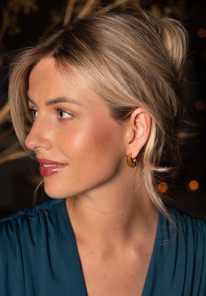 Gold-tone hoop earrings with a textured, organic shape. Worn by a person with light blonde hair, soft features, and a teal garment.