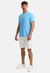 Helles blaues T-Shirt mit kurzen Ärmeln und strukturiertem Design, ausgestattet mit einer Brusttasche, kombiniert mit weißen, hochgekrempelten Shorts und weißen Sneakern.