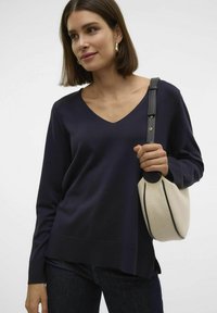 Pull bleu marine en V à manches longues, avec un ourlet côtelé, associé à un sac crème avec une bordure noire. Le tissu semble doux et texturé.