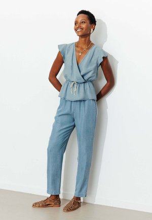 Femme souriante en haut cache-cœur sans manches bleu clair et pantalon assorti, debout les mains dans le dos, portant des sandales à imprimé léopard devant un mur blanc.