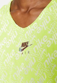 Neon zelený top Nike s výstřihem do lodi, opakujícím se textovým vzorem a stříbrným logem Nike s nápisem "AIR" pod ním ve kontrastní barvě.