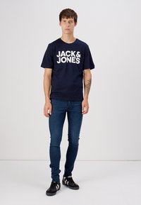 Jūrų mėlynos medvilnės marškinėliai su dideliu baltu tekstu "JACK & JONES". Suderinta su tamsiomis džinsinėmis kelnėmis. Modelis dėvi juodus batus su baltais dryžiais.
