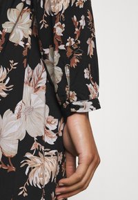 Schwarzes Blumenkleid mit großen beigen und braunen Blumen, aus strukturiertem Stoff und mit Knopfdetails an den Ärmeln.