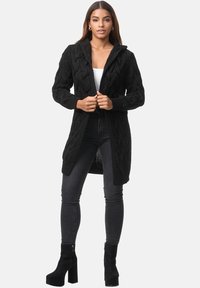 Cardigan nero a maglia lavorata a trecce con cappuccio, dotato di maniche lunghe, chiusura con bottoni e taglio a metà lunghezza, abbinato a jeans scuri e stivali con tacco.