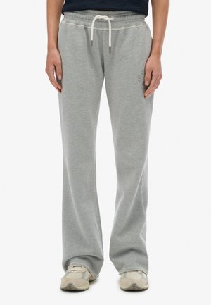 Persoon in lichtgrijze sweatpants met een witte koord en beige sneakers, staande tegen een effen witte achtergrond.